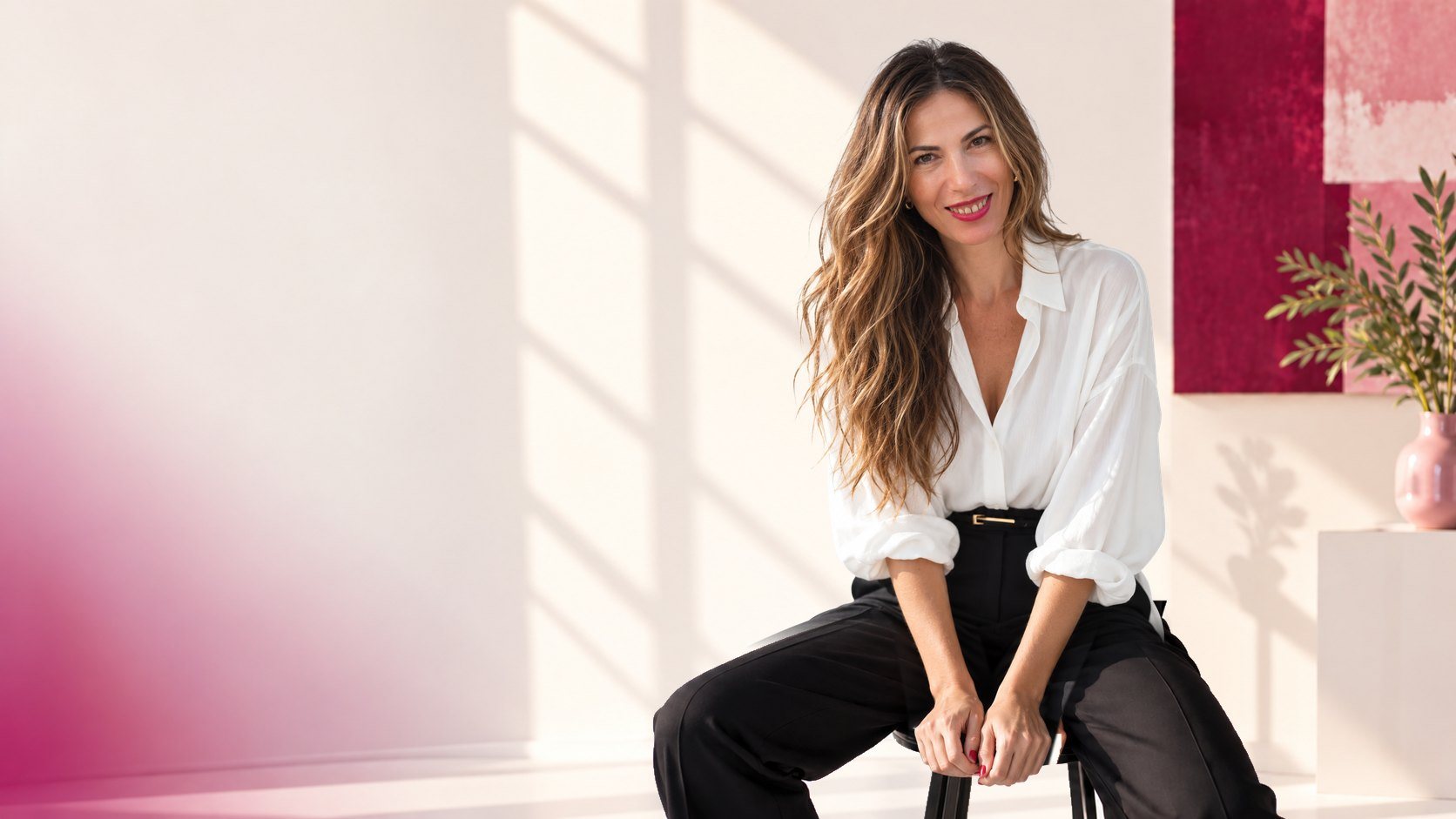 Sara con camisa blanca y pantalón negro, sentada en un taburete en un estudio luminoso, sonriendo a cámara.