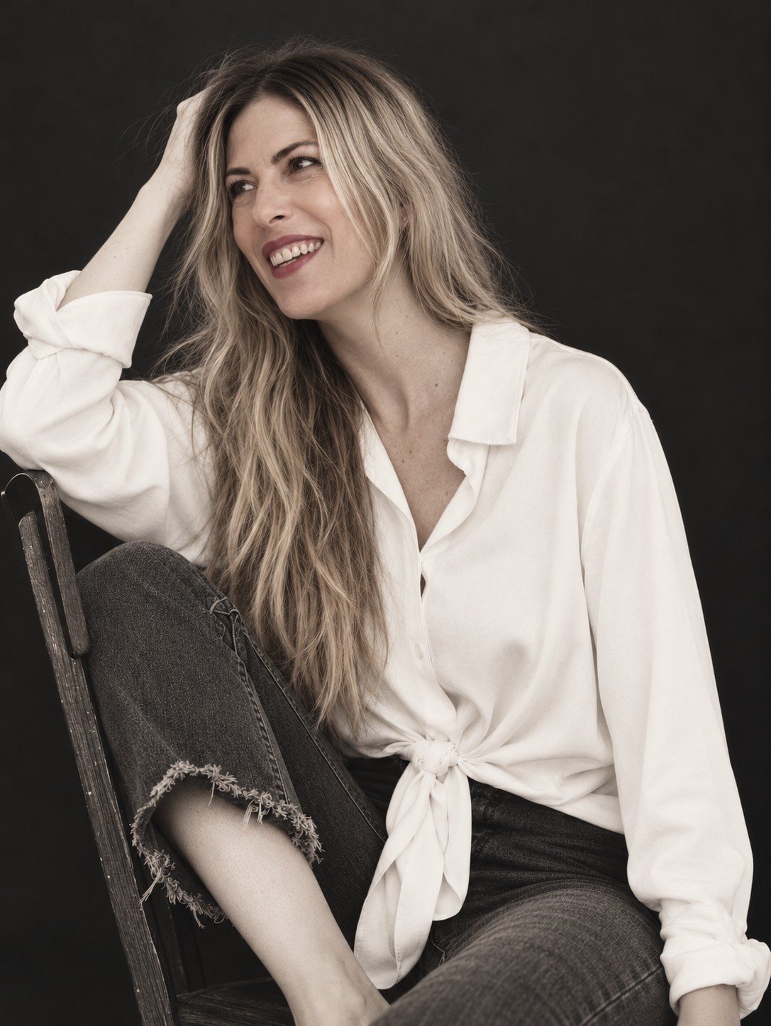 Retrato de Sara sonriendo, con camisa blanca anudada, sentada en una silla de madera sobre fondo oscuro.